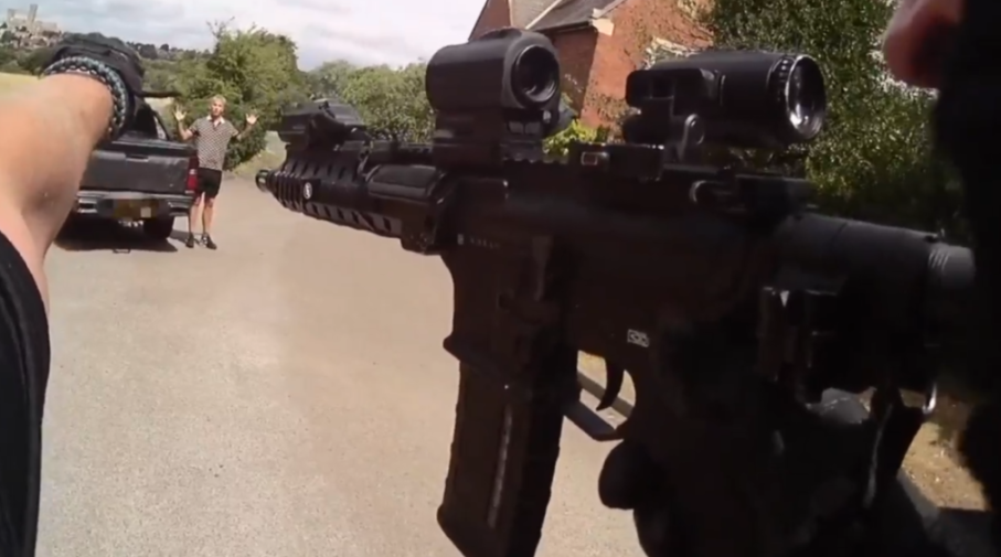 A person with an assault rifle pointed at a man standing with his hands up behind a pickup truck.