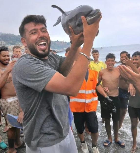 A man holds up a dolphin on a beach surrounded by other people.