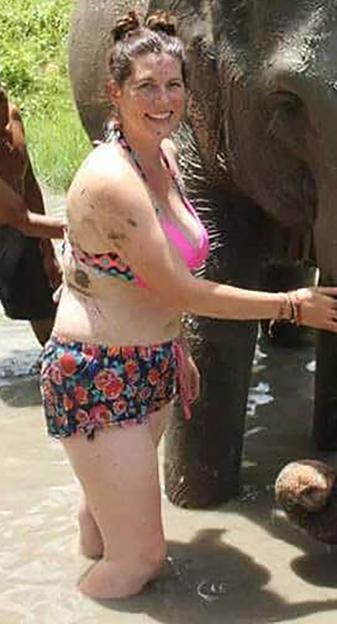 A smiling woman in a bikini and floral shorts stands in shallow water, next to an elephant.