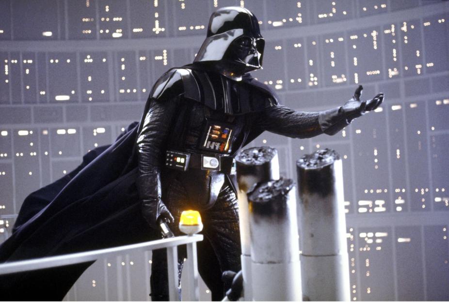 Darth Vader, in his iconic black suit and helmet, stands on a platform with his arm outstretched, against a background of glowing lights.