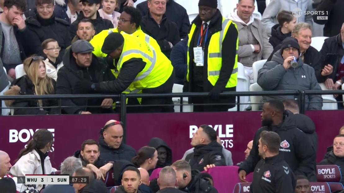 A man in a flat cap speaks with a security guard in a yellow vest, while other people watch a game.