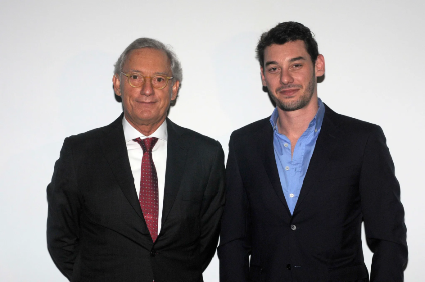 Two men in suits standing side-by-side, the man on the left older, the man on the right younger.