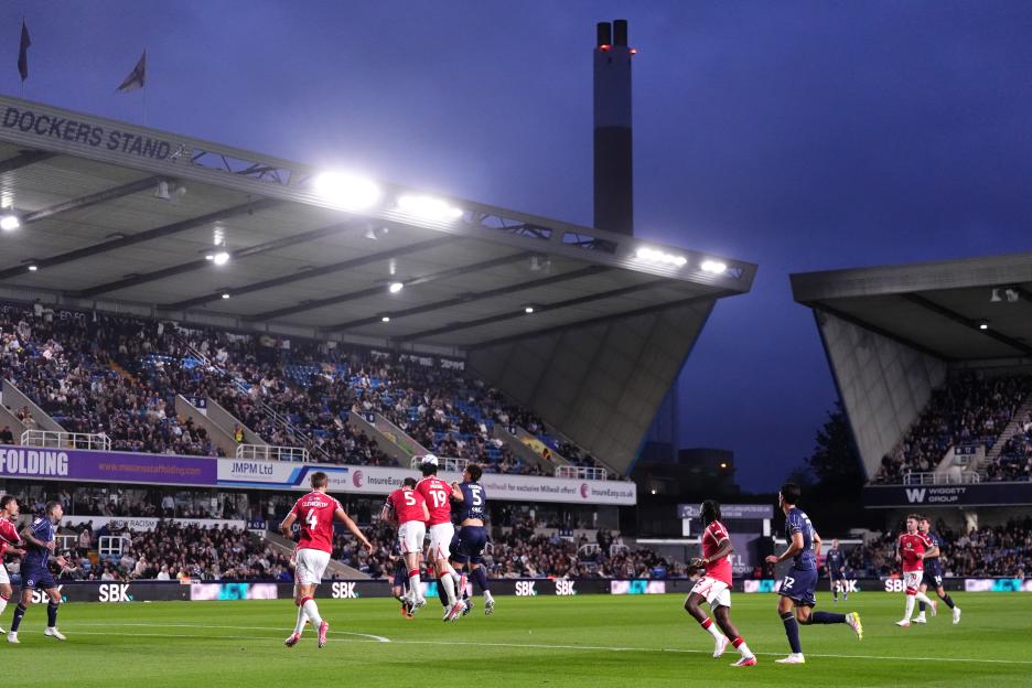 A wide shot of a soccer match in progress with players in red and blue uniforms on the field and spectators in the stands, under a twilight sky.