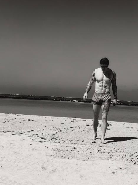 Black and white image of Adam Peaty walking on a beach, revealing a new tattoo on his abdomen.