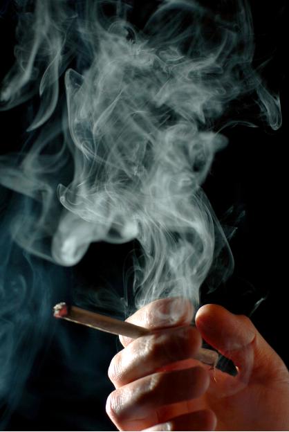 A hand holding a lit cigarette with smoke rising from it against a dark background.