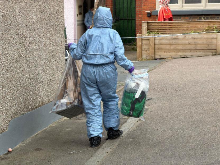 A police officer in a blue forensic suit and purple gloves carries two clear evidence bags.