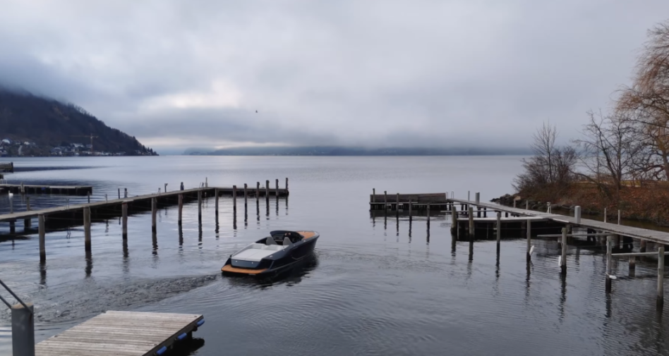 A modern boat navigating between wooden docks on a lake, with a cloudy sky and mountains in the background.
