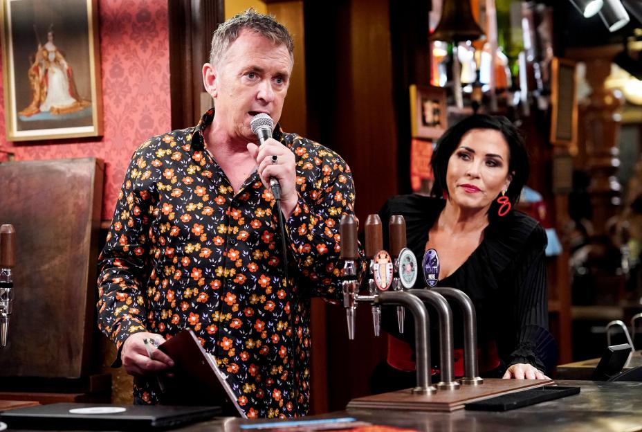 A man in a floral shirt speaks into a microphone as a woman watches behind a bar.