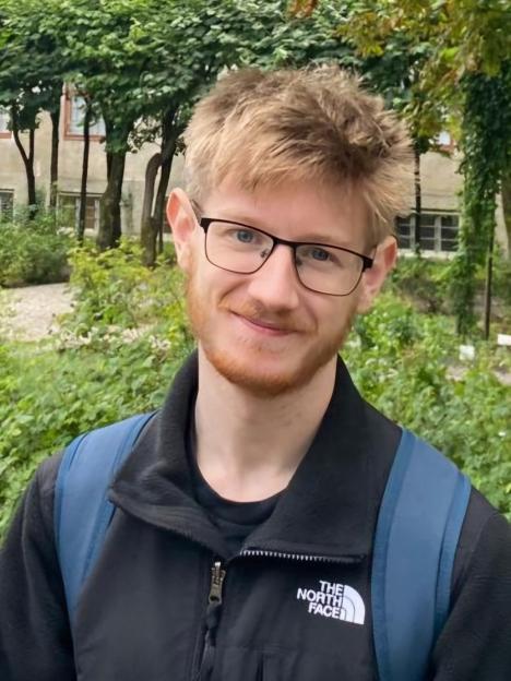 Fintan Jack Feltham, an 18-year-old student, smiling and wearing glasses, a black jacket, and a blue backpack.