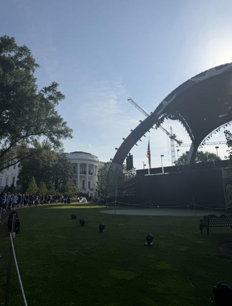 The White House with an outdoor stage and large cranes visible in the background, as people gather on the lawn.
