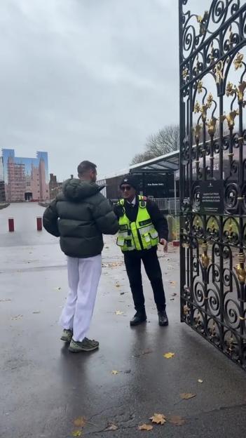 A man in a green puffer jacket and white pants speaks to a security guard in a high-visibility vest next to an ornate black and gold gate. In the background, a building resembling a castle is visible.