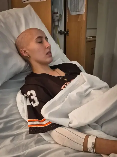 A young, bald woman with closed eyes lying in a hospital bed, wearing a dark brown and orange sports jersey.