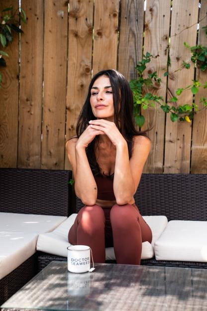 A woman sitting on an outdoor sofa with a mug labeled "DOCTOR SEAWEED" on the table.