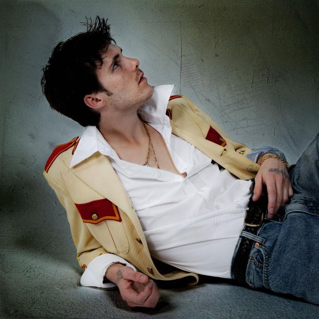 Cruz Beckham in a cream and red jacket, unbuttoned white shirt, and jeans, lying down and looking up.