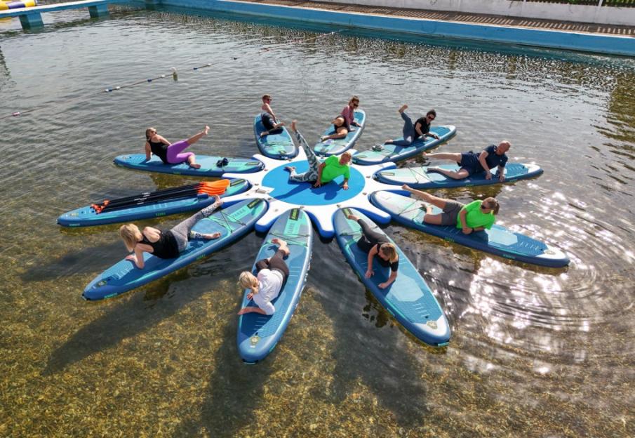 People in a paddle boarding fitness class doing exercises on boards arranged around a central mat in the water.