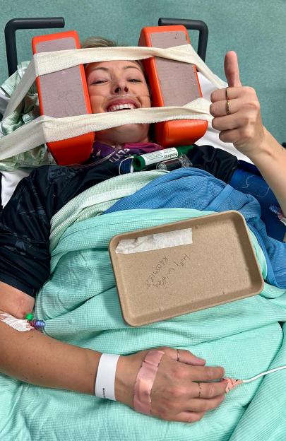 A woman in a neck brace and nasal cannula gives a thumbs-up from a hospital bed.