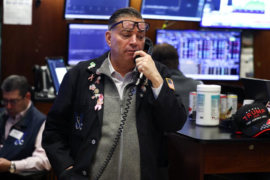 A man in glasses wearing a grey sweater, a black jacket covered in pins, and a "Trump 2024" hat nearby, speaks into a phone while looking at stock market screens.