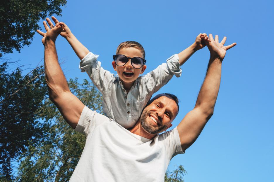 Father and 5 years old son having fun outdoors, holding a boy up against blue sky. Boy has large funny sunglasses on