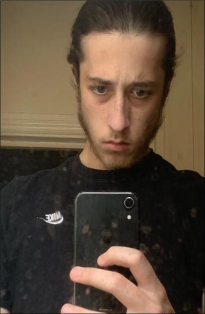 Alfie Coleman, a man with long dark hair pulled back, looks directly at the camera while holding a black smartphone to take a selfie.