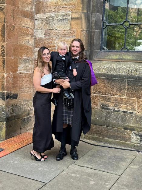A man in graduation robes and kilt holds a young child in a suit and kilt, standing next to a woman in a black and white dress.