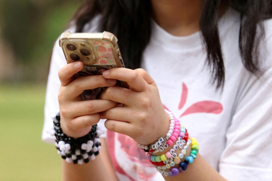 A girl holds a smartphone with two hands, wearing multiple colorful beaded bracelets on her left arm.