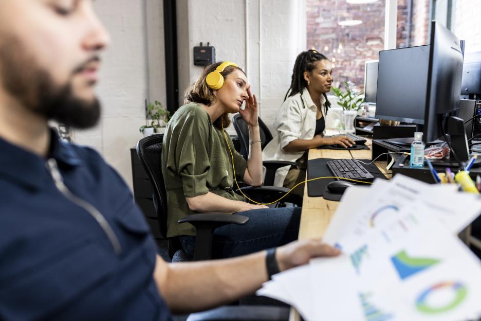 A frustrated businesswoman with yellow headphones at a desk with other coworkers.