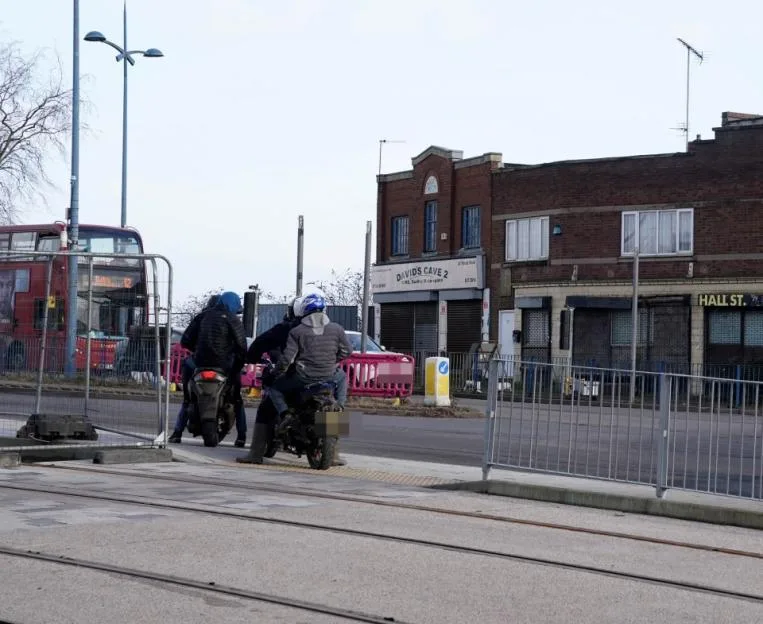 Two people on mopeds stopped on a street corner, with a double-decker bus and buildings in the background.