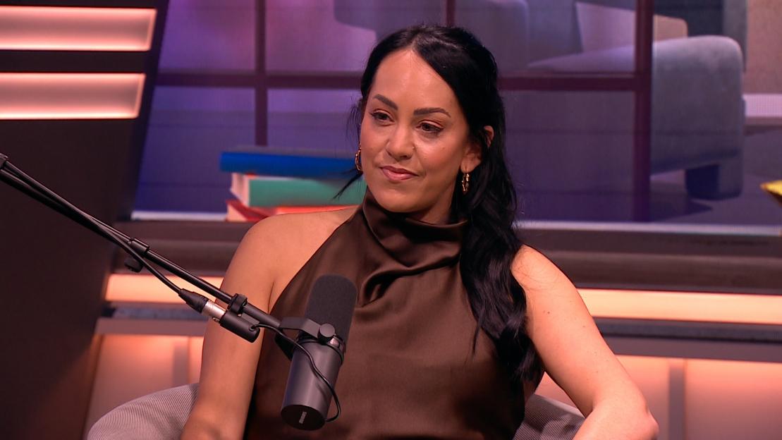 A woman with dark hair wearing a brown halter-neck top speaks into a microphone.