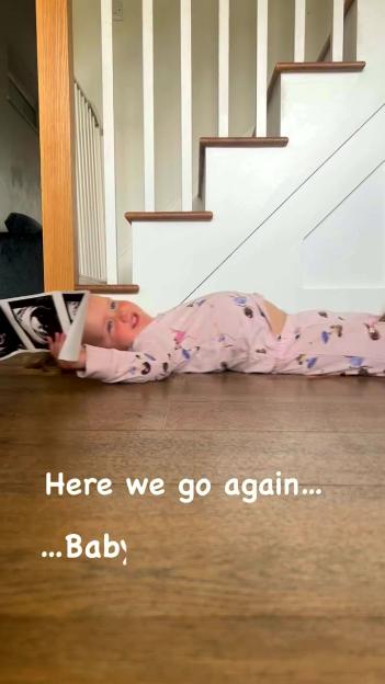 A child in pink pajamas lying on the floor, holding ultrasound photos, with text "Here we go again... ...Baby" signaling a pregnancy announcement.