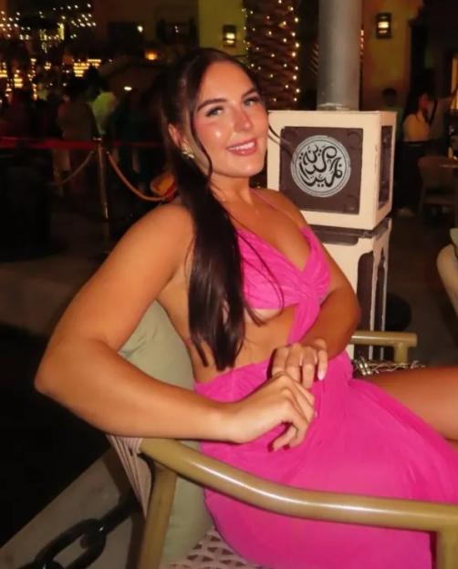 A woman in a pink dress smiles at the camera while sitting in a restaurant at night.