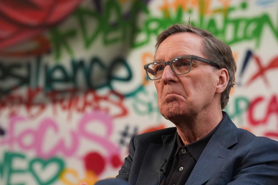 Former health secretary Alan Milburn with a concerned expression, wearing glasses and a dark suit, against a colorful, graffiti-covered wall.