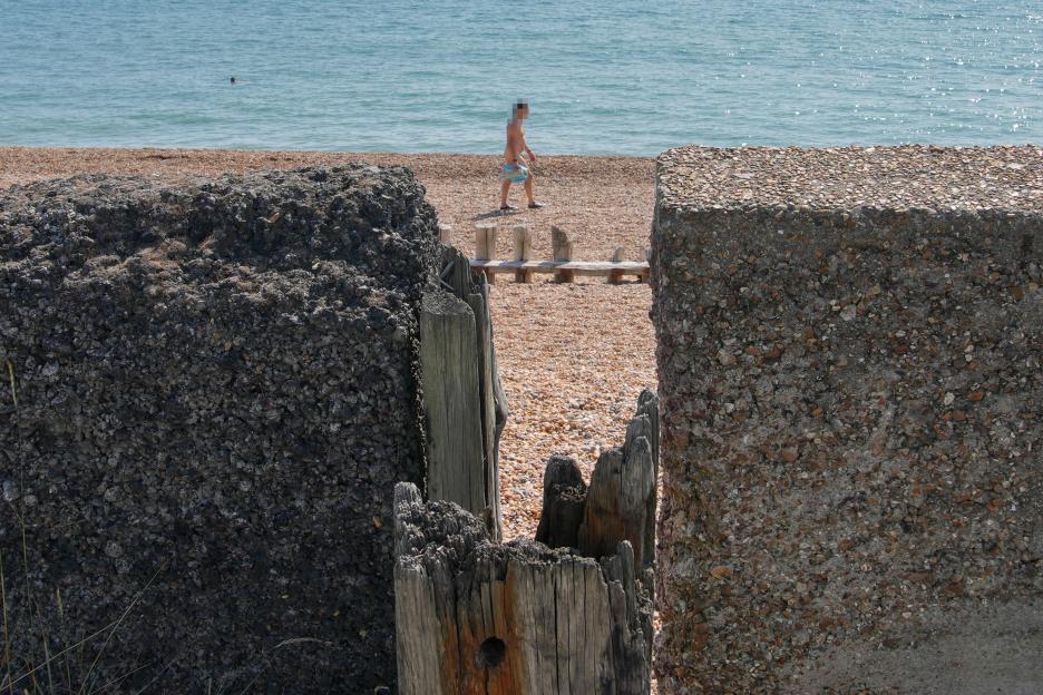 A nudist walking on a pebble beach, seen through a gap in a concrete wall.