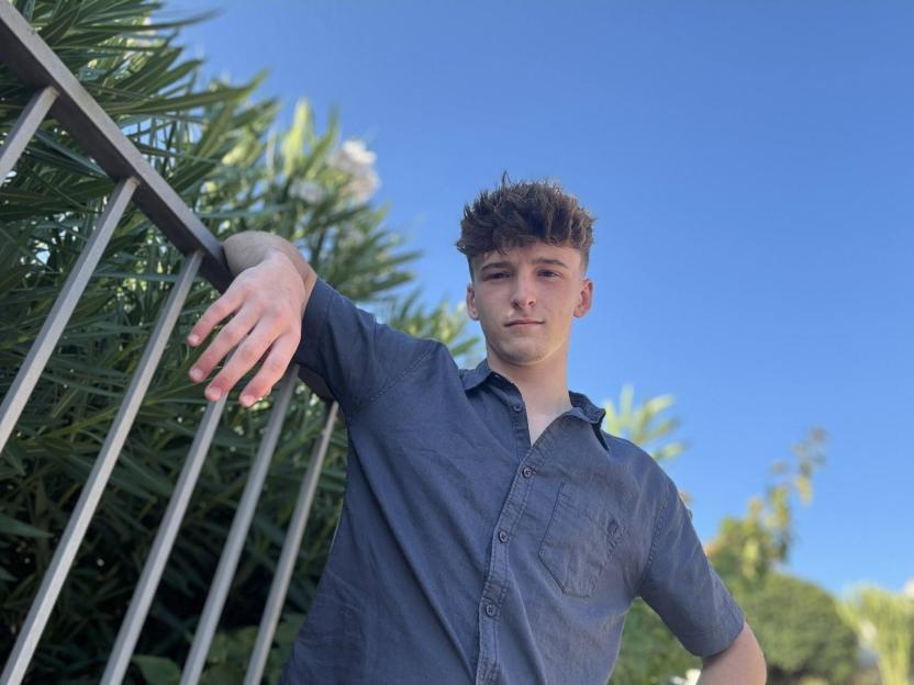 A young man with spiked brown hair and a dark blue shirt leans on a metal railing with a blue sky and green foliage in the background.