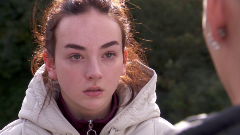 A young woman with brown hair, in a cream colored puffer coat with a purple shirt underneath, looking at a person to her right with an expression of concern.