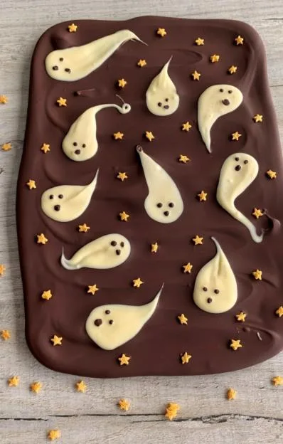 A slab of dark chocolate bark decorated with white chocolate ghosts and orange star sprinkles.