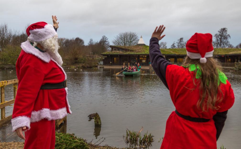Santa and an elf waving at children in a boat on a lake.