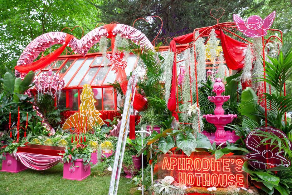 The "Aphrodite's Hothouse" garden exhibit features a greenhouse decorated with red and pink fabrics, hearts, cupids, and lush greenery, including a neon sign that reads "Aphrodite's Hothouse."
