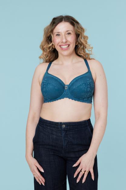 A smiling woman with curly hair wears a teal bra and dark jeans against a light blue background.