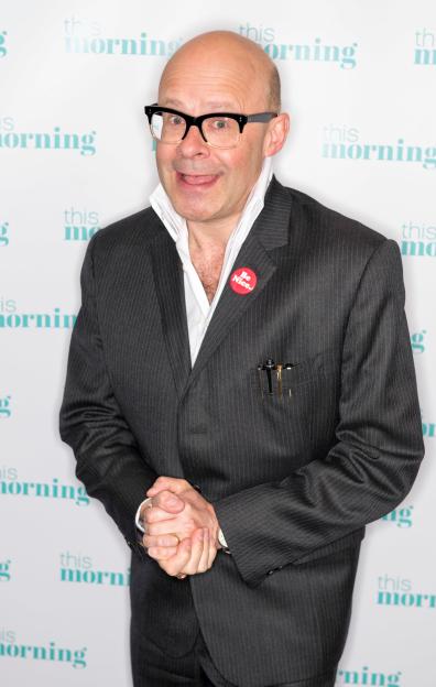 Comedian Harry Hill wearing a pinstripe suit, white shirt, black-rimmed glasses, and a "Be Nice" button.