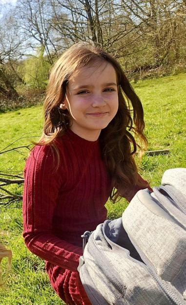 Aria Thorpe, a young girl with long brown hair, sitting on grass and smiling.