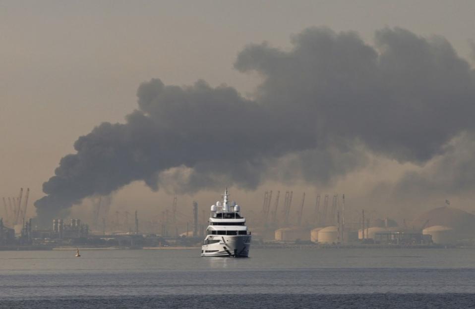 A large white yacht floats on water in front of a hazy industrial landscape with plumes of dark smoke rising into the sky.