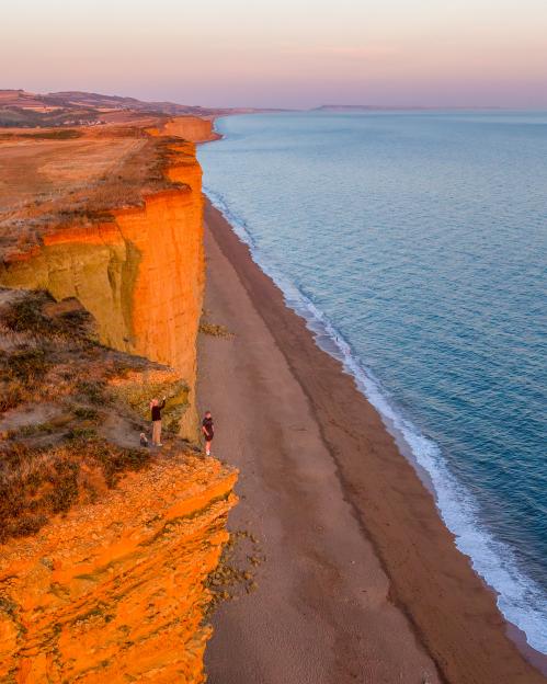 Two people standing on the edge of a crumbling 150ft cliff overlooking a beach and ocean.