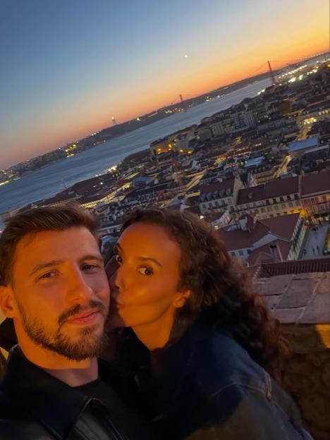 Maya Jama kissing Ruben Dias on the cheek with a city and river at sunset in the background.