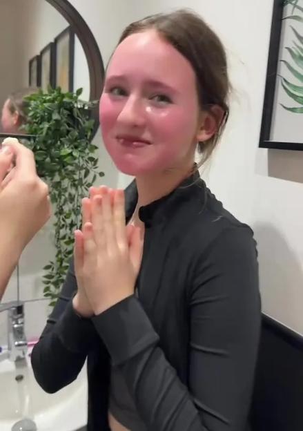 A girl with a pink, stained face, wearing a black shirt, puts her hands together at the sink.