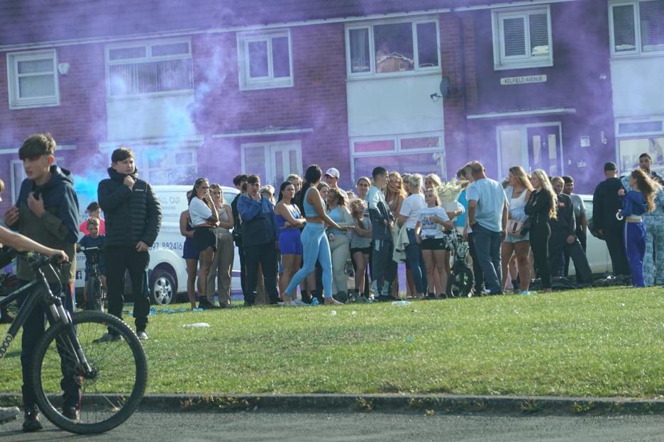 A large group of people stand on a grassy area in front of brick buildings with purple smoke filling the air.