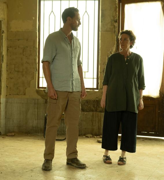 Three individuals, a soldier in camouflage, a man in a light-colored shirt and khaki pants, and a woman in a dark green top and black pants, stand inside a dilapidated building.
