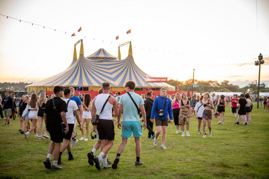 Festival attendees walking on grass with a large striped tent in the background.