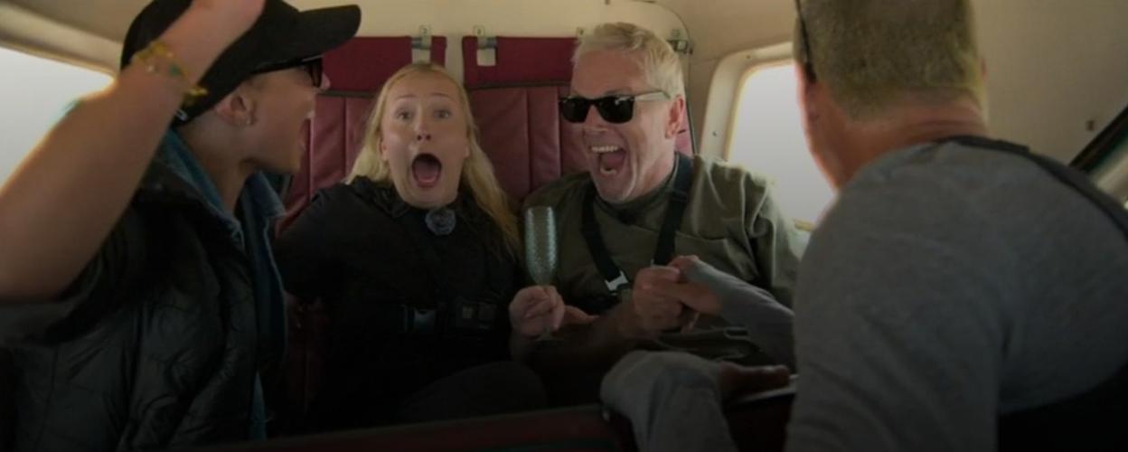 Three people with excited expressions in what appears to be a small plane, with one person holding a champagne flute.