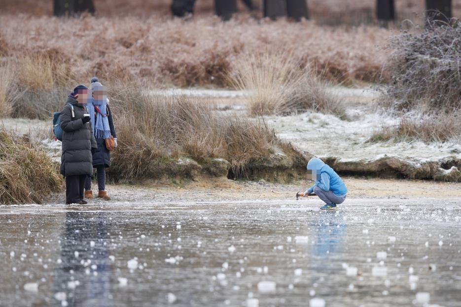 A child squats on a frozen pond with a hammer while two adults stand nearby.