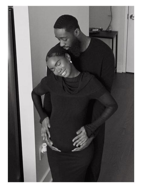 Black and white photo of a man embracing a pregnant woman from behind, her hands cradling her belly.